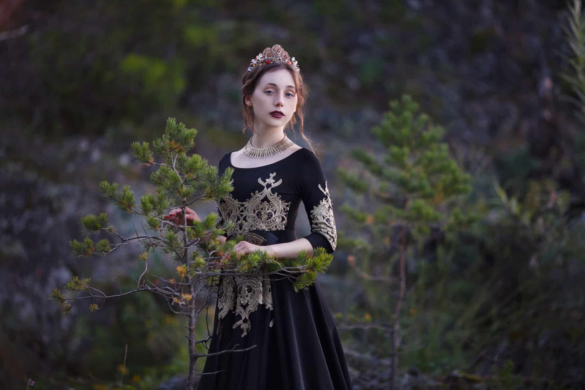 sad young woman in a black dress with a crown on her head stands on the banks of the river