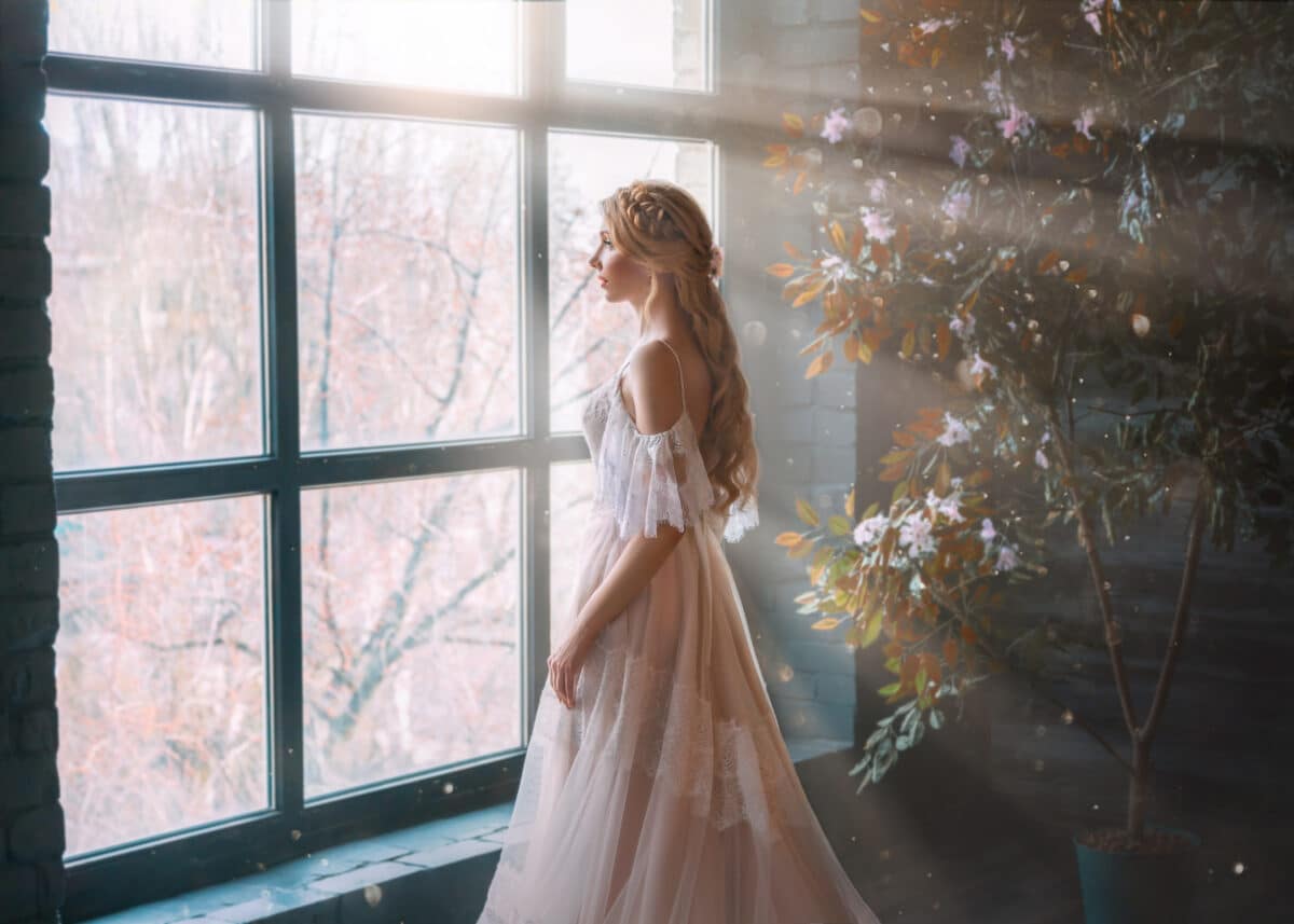 Romantic noble lady with long hair in white vintage dress stands in dark room, looks out window