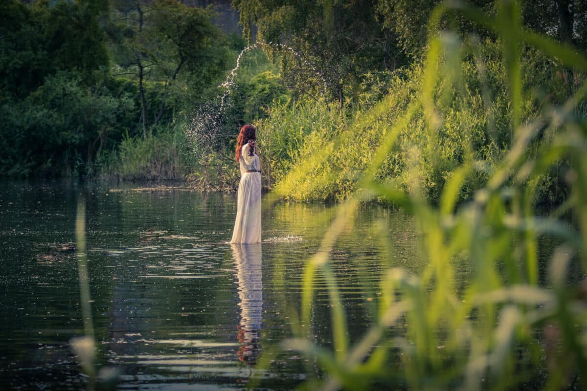 nymph with long red hair in a white dress walking in the river