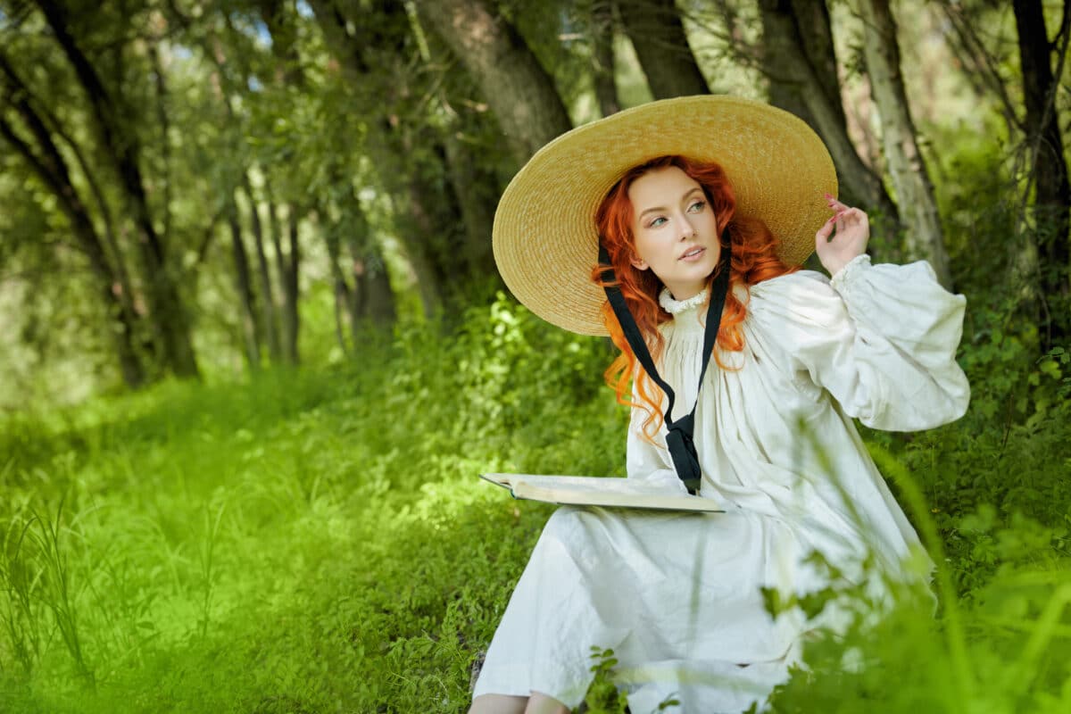 A romantic red haired lady with charming wavy red hair, dressed in a long white shirt and a wide-brimmed straw hat, sits with a book in a park on a sunny summer day