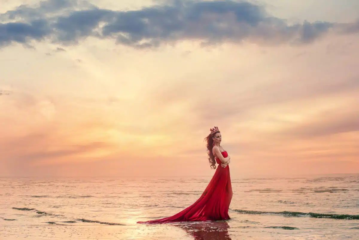 Beautiful woman in fluttering red dress on sea wave at sunset.
