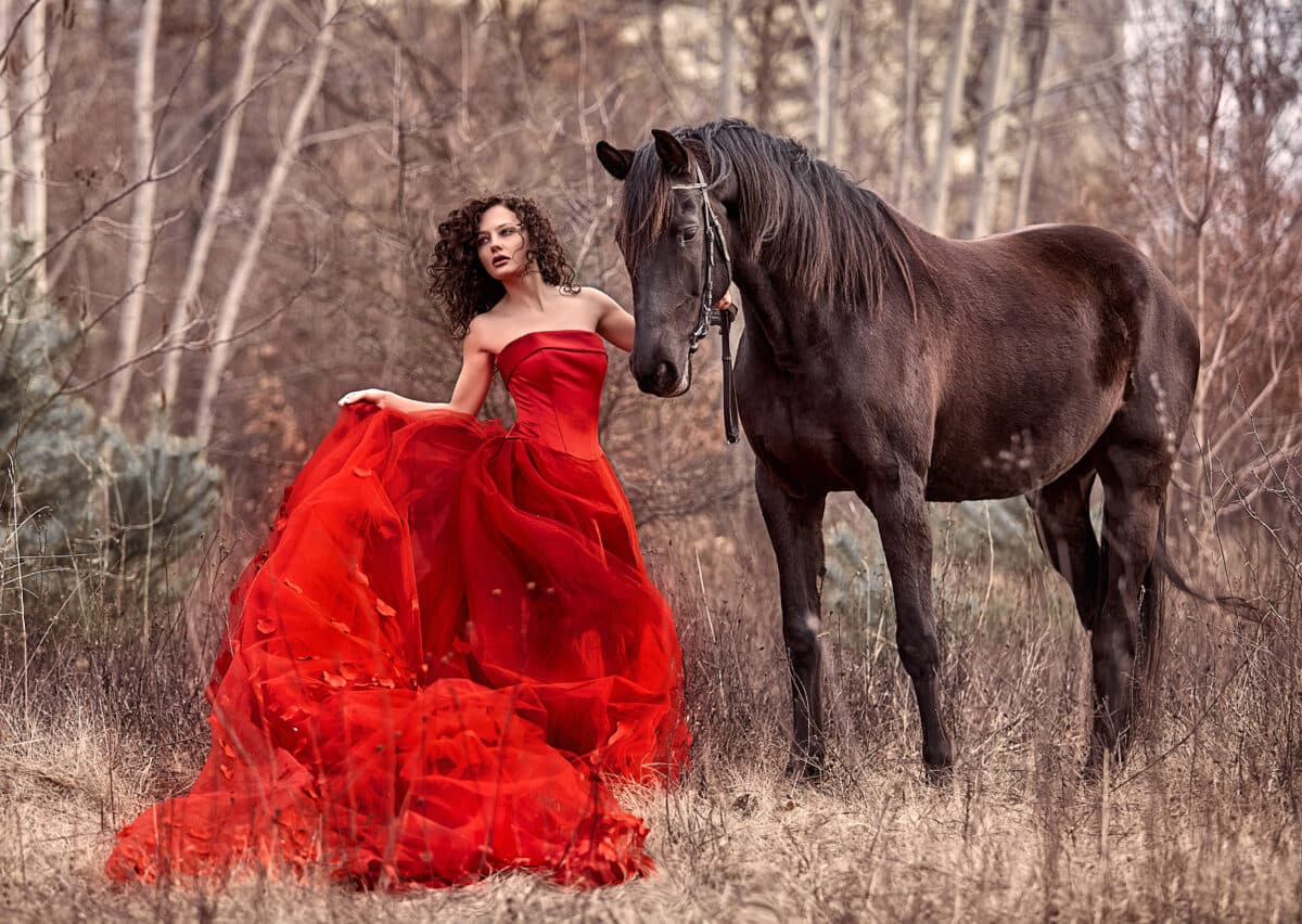 A beautiful lady in a red dress stands next to a brown horse in the woods