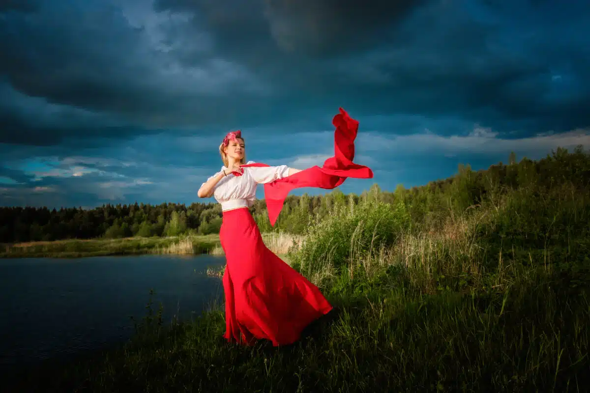 A slender woman in a red skirt and white blouse is dancing with a red shawl in her hands against a stormy sky