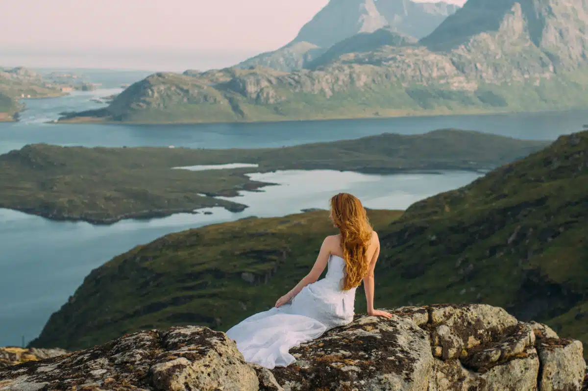 Back of girl in wedding dress sitting on rocks in mountains and looking to fjord