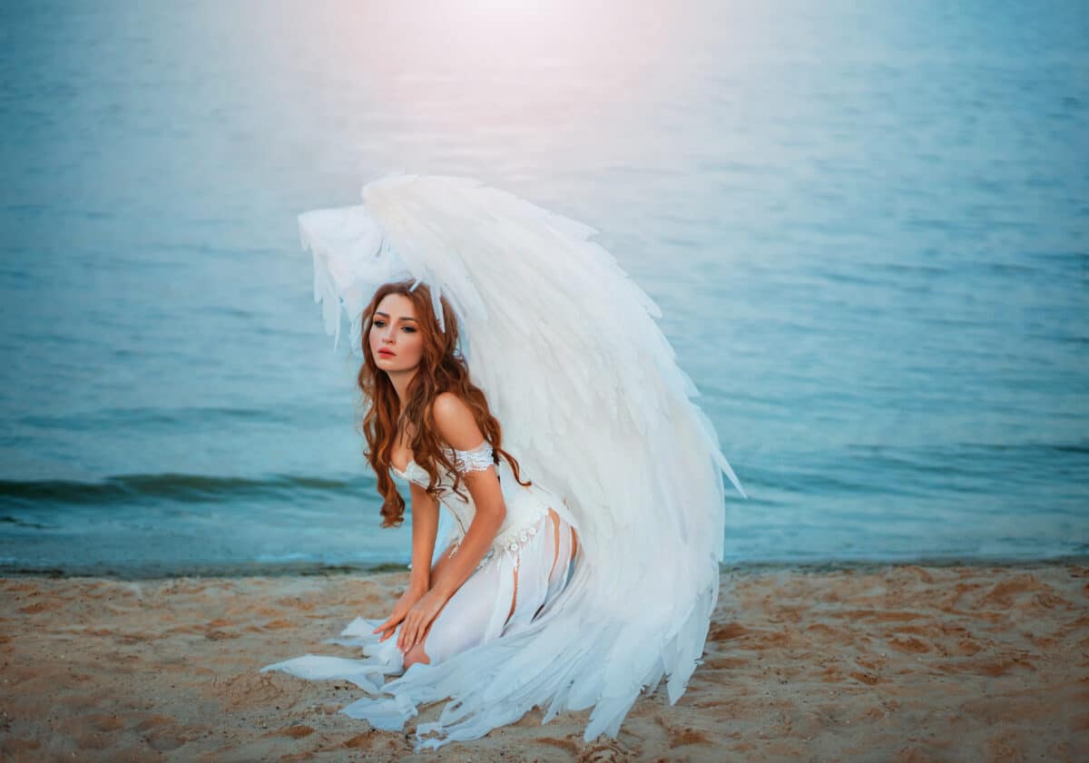 Young beautiful woman fallen sad angel sits on the sea beach. Creative sexy costume, huge artificial bird wings and white vintage dress. Adult girl with sad face of repentance. Artwork photo