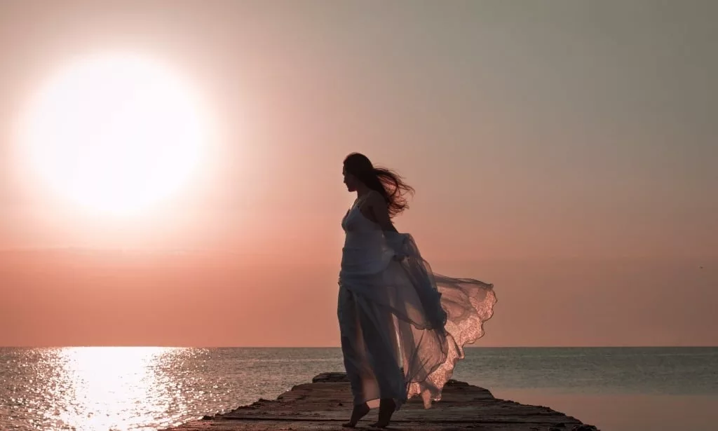 Woman in flowy dress standing by the sea and sunset.