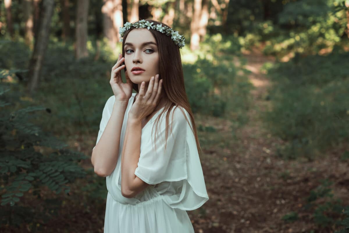beautiful tender girl posing in white dress and floral wreath in forest