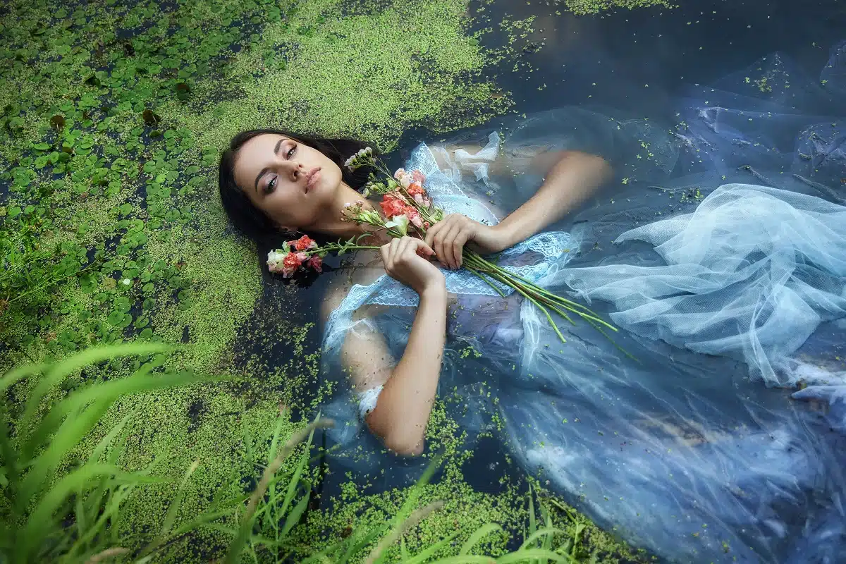 enchanting woman lies in river in blue long dress with flowers