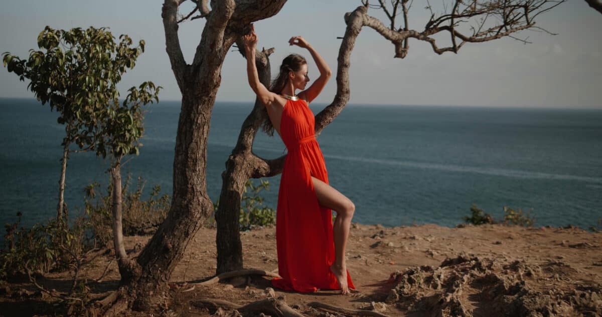 Beautiful woman in red dress standing on the sea shore cliff during sunny summer day