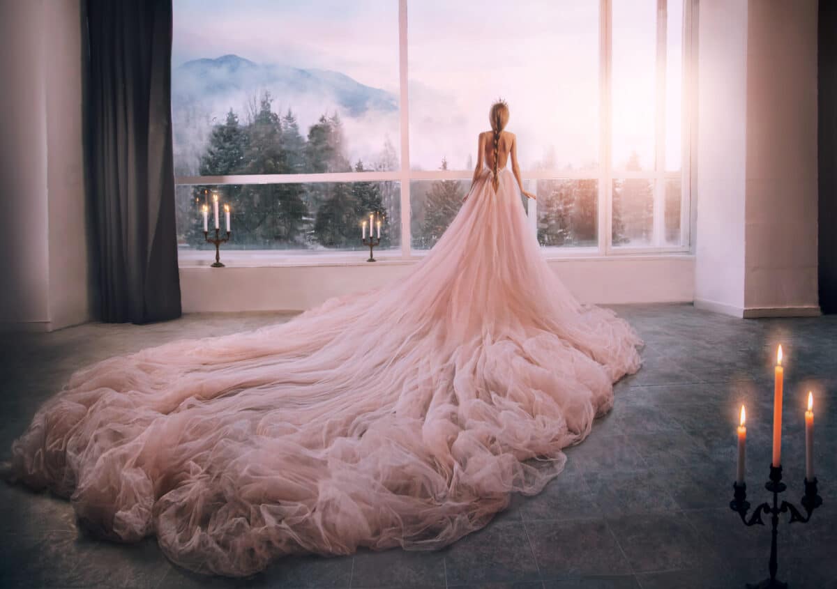 Fantasy girl princess in pink dress stands in medieval castle room looking vintage window with winter nature landscape mountains sunset. Mysterious silhouette woman queen, long train skirt. Back view