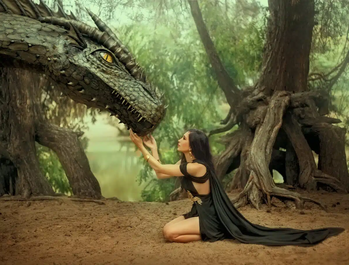 Fantasy woman elf queen touching with hands dragon head. Girl mistress tamed monster concept female power. black creative dress, girl princess fashion model sits on knees. Deep green forest trees