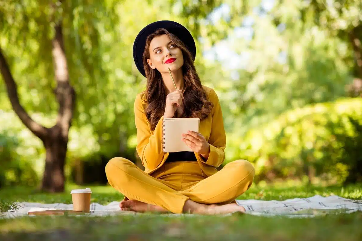 Charming gorgeous wavy-haired girl sitting in lotus position writing outdoors.