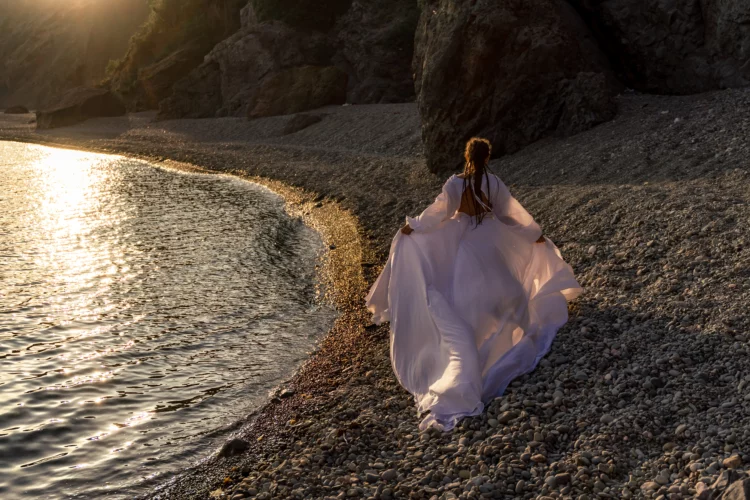 A mysterious woman in long dress with long braids running on the sea beach with mountain views at sunset