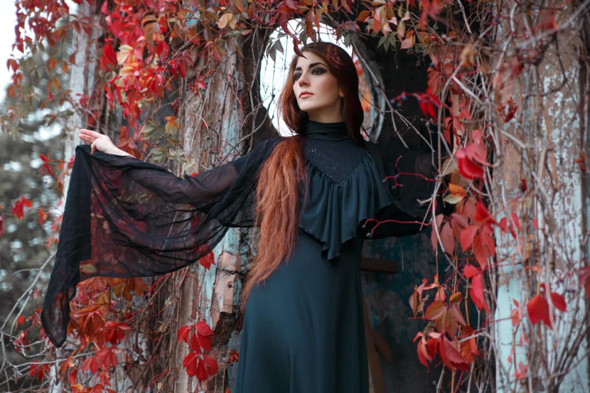 Portrait of a beautiful mysterious woman with long red hair in a black dress on the background of an old building entwined with red autumn vine leaves.