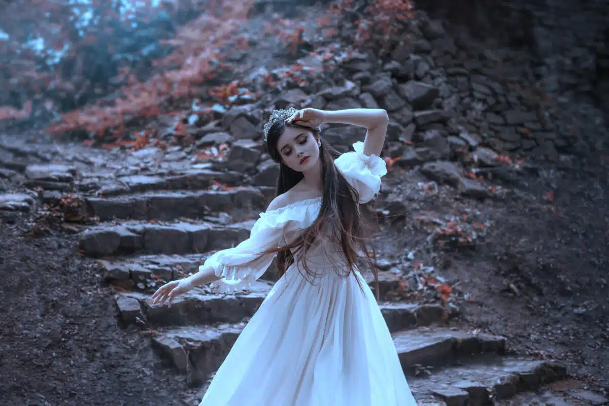 Young princess with very long hair posing against the background of an old stone staircase. The girl has a crystal crown and a white, flying vintage dress. Artistic processing, unusual colors