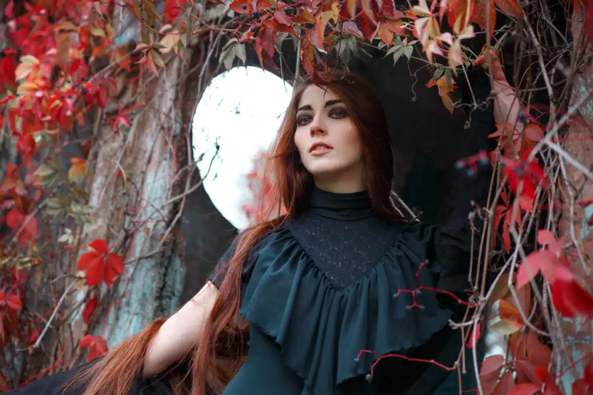 Portrait of a beautiful mysterious woman with long red hair in a black dress on the background of an old building entwined with red autumn vine leaves.