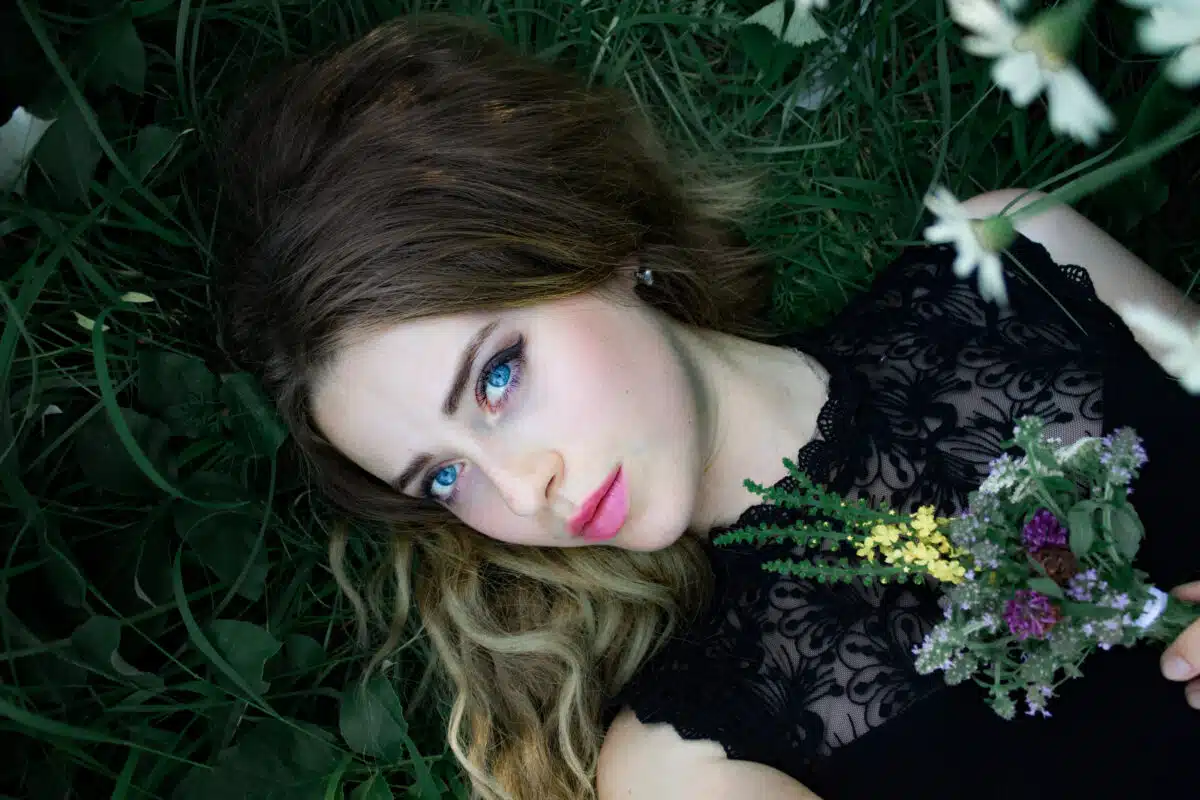 A woman with blue eyes lies on the grass with a bouquet of wildflowers in her hands.