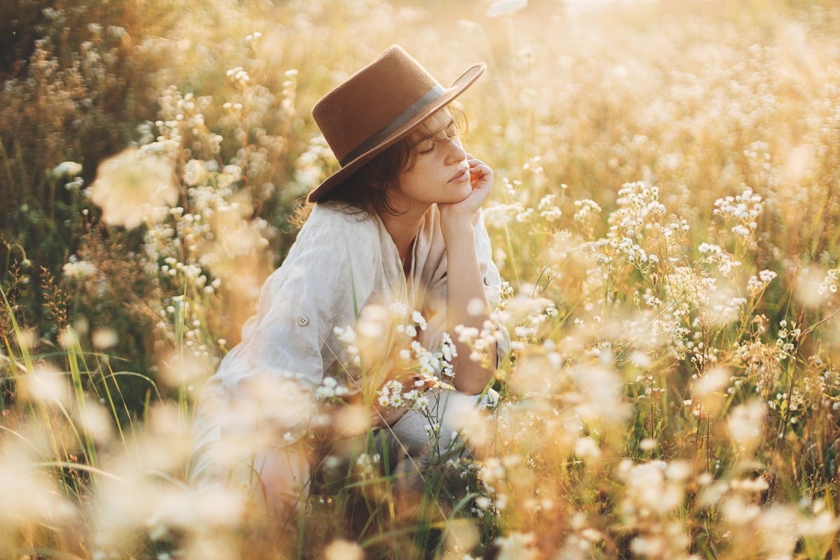 boho woman in hat sitting among wildflowers in warm sunshine