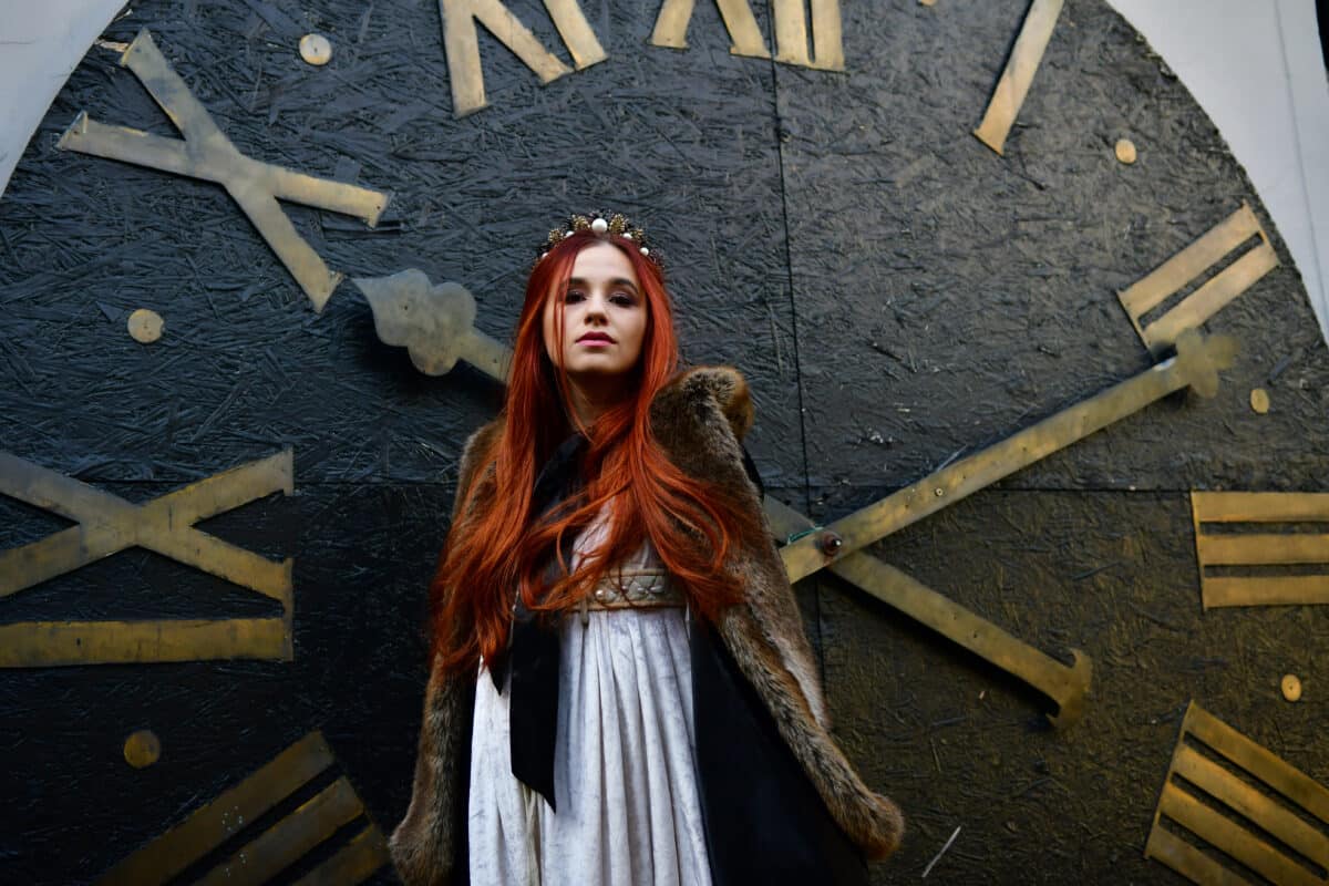 Red-haired princess in a white dress and a black veil with a crown on her head near the large clock face with hands