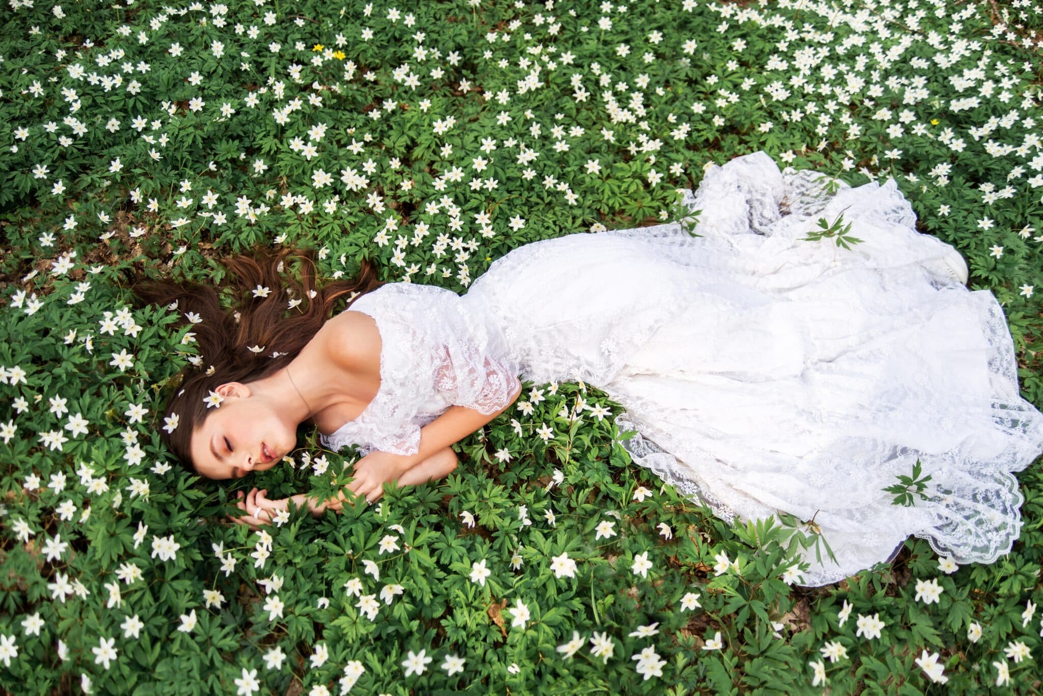 girl in a white lace dress lies in a clearing with primroses. A young woman with long hair is sleeping in a spring forest.