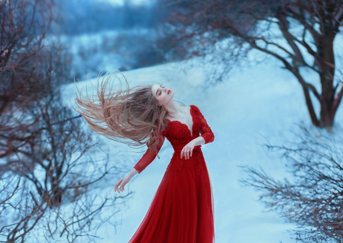 A young girl with very long hair flying in the wind. Princess in a red dress on a background of winter hills and gloomy trees. Cold colors, beautiful winter.