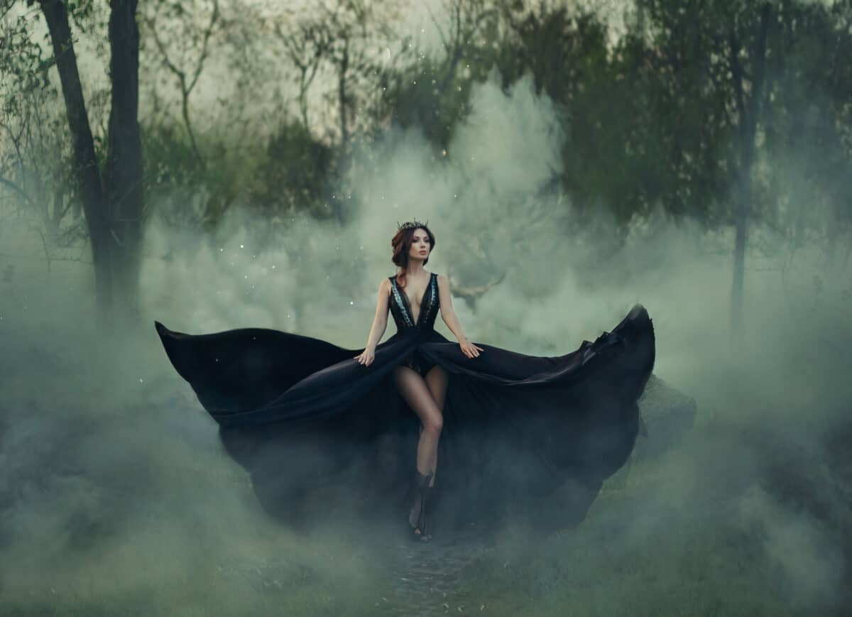 Fantasy gothic woman dark queen bare long legs walks in fog. black dress fly in different directions, like wings of raven bird. Elegant hairstyle crown. Artistic photo. Deep dark forest woods trees