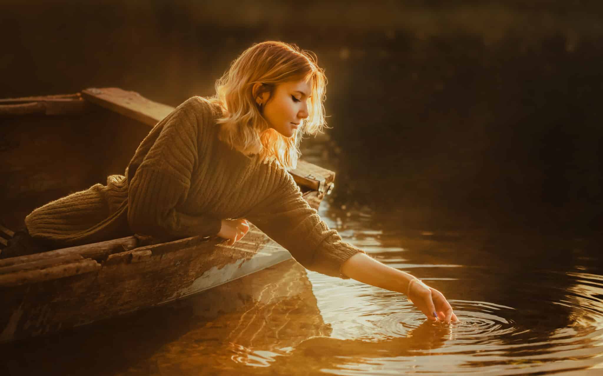 A beautiful blonde woman sits in an old fishing boat reaches and touches the water in the lake