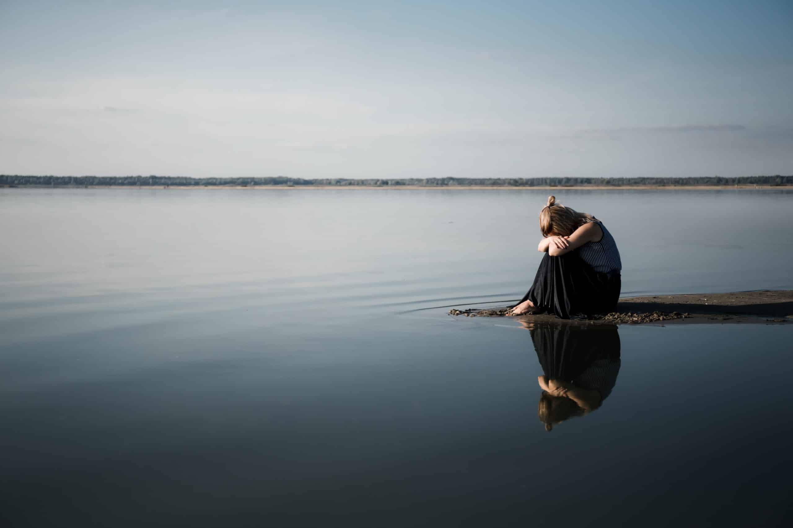 A young woman sits on the shore of the lake with her head on her knees.