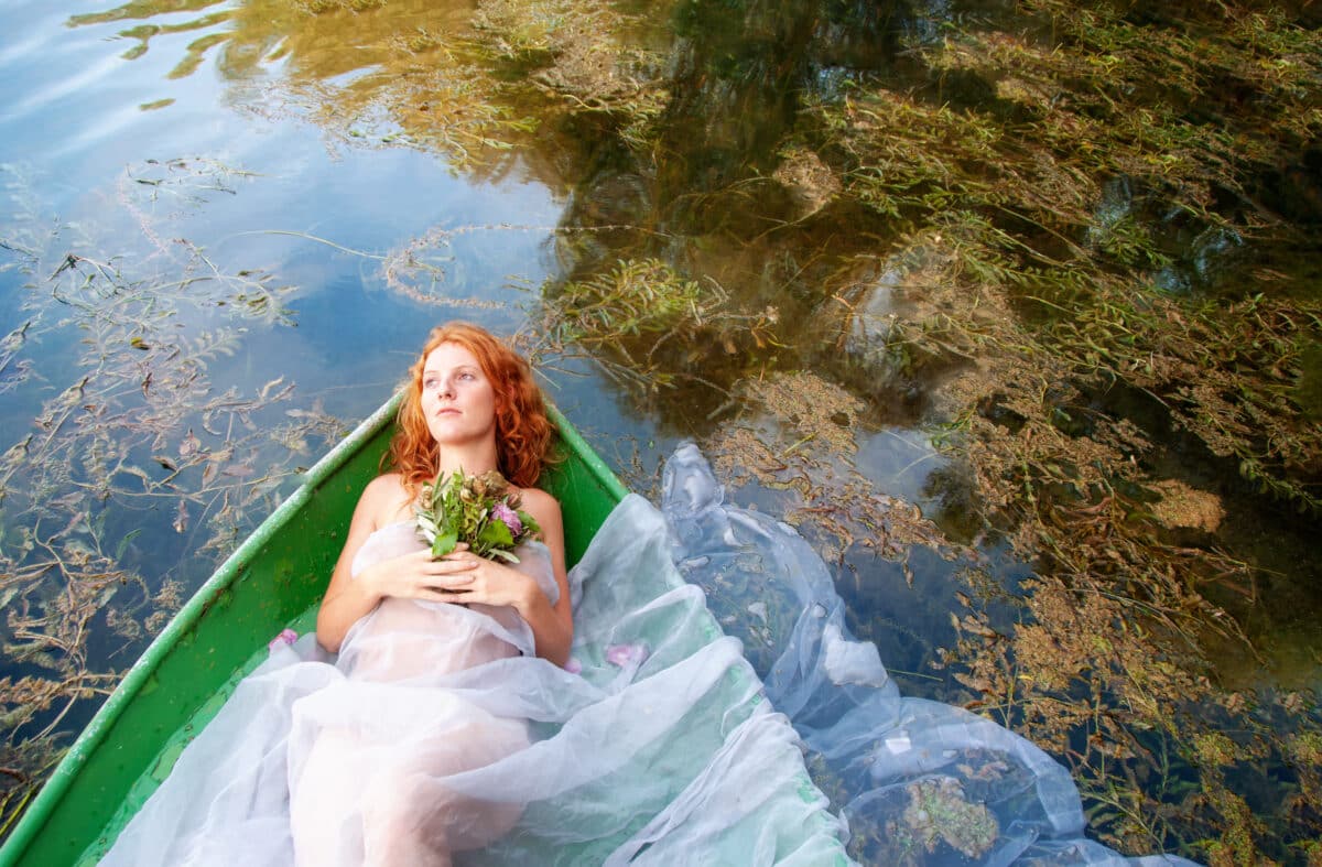 beautiful sensual, seductive, young sexy redhead woman with a longing, bride, Lady of Shalott, with a bouquet of wilted flowers lying in a boat in lake water