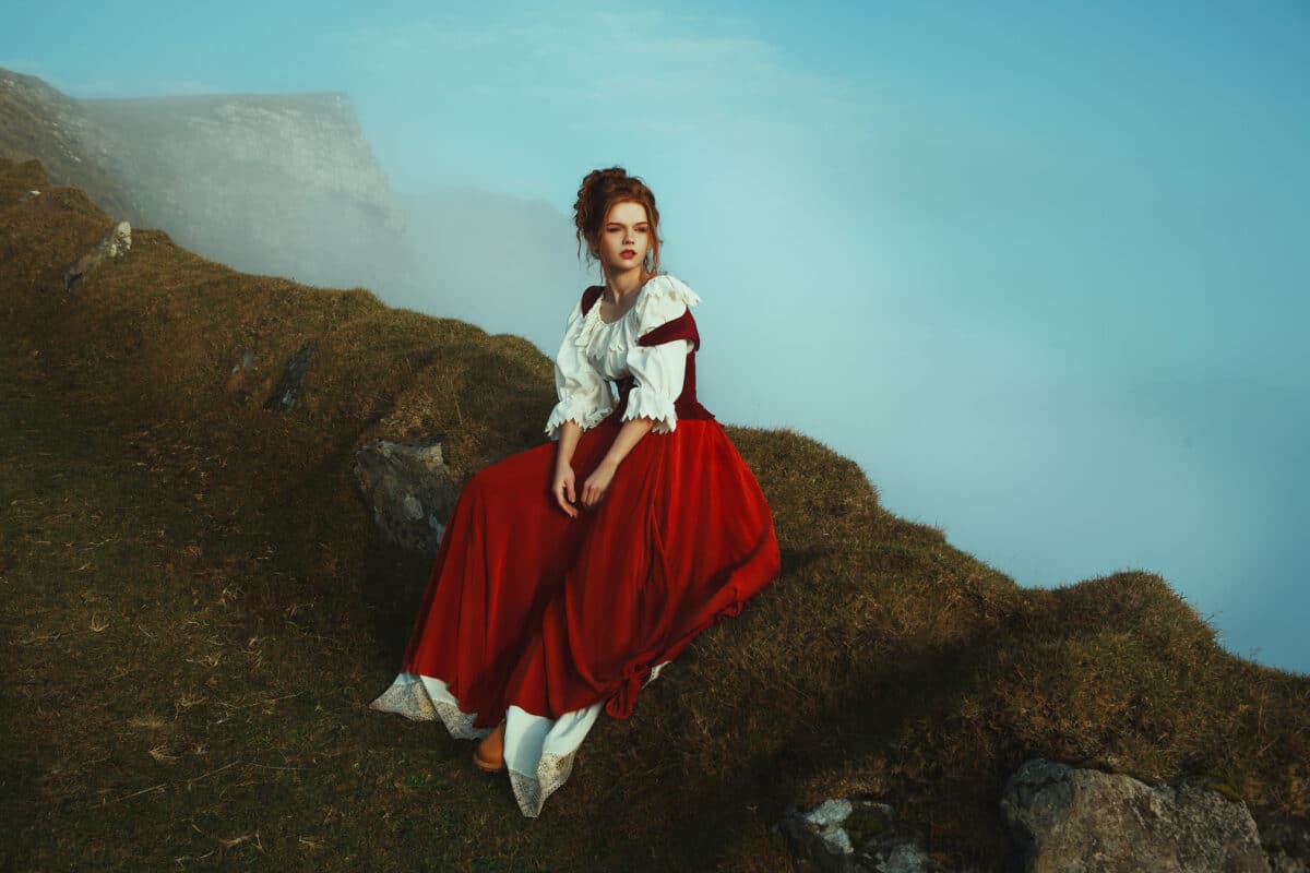 Beautiful red-haired girl is sitting on the stones covered with moss, over the precipice at the background of mountains and mist
