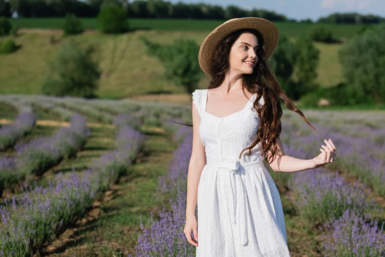 Happy woman in white dress and straw hat looking away in meadow with blooming lavender.