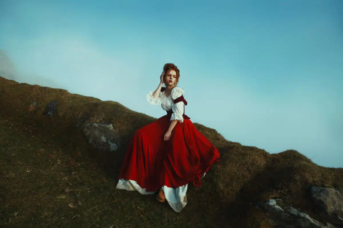 Beautiful red-haired girl is sitting on the stones covered with moss, over the precipice at the background of mountains and mist