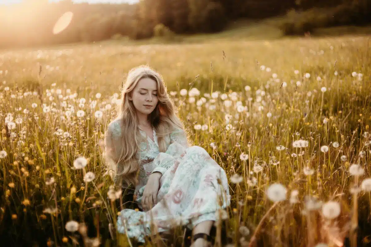 a romantic young blonde lady in summer sits in a field of dandelions at sunset in a long dress