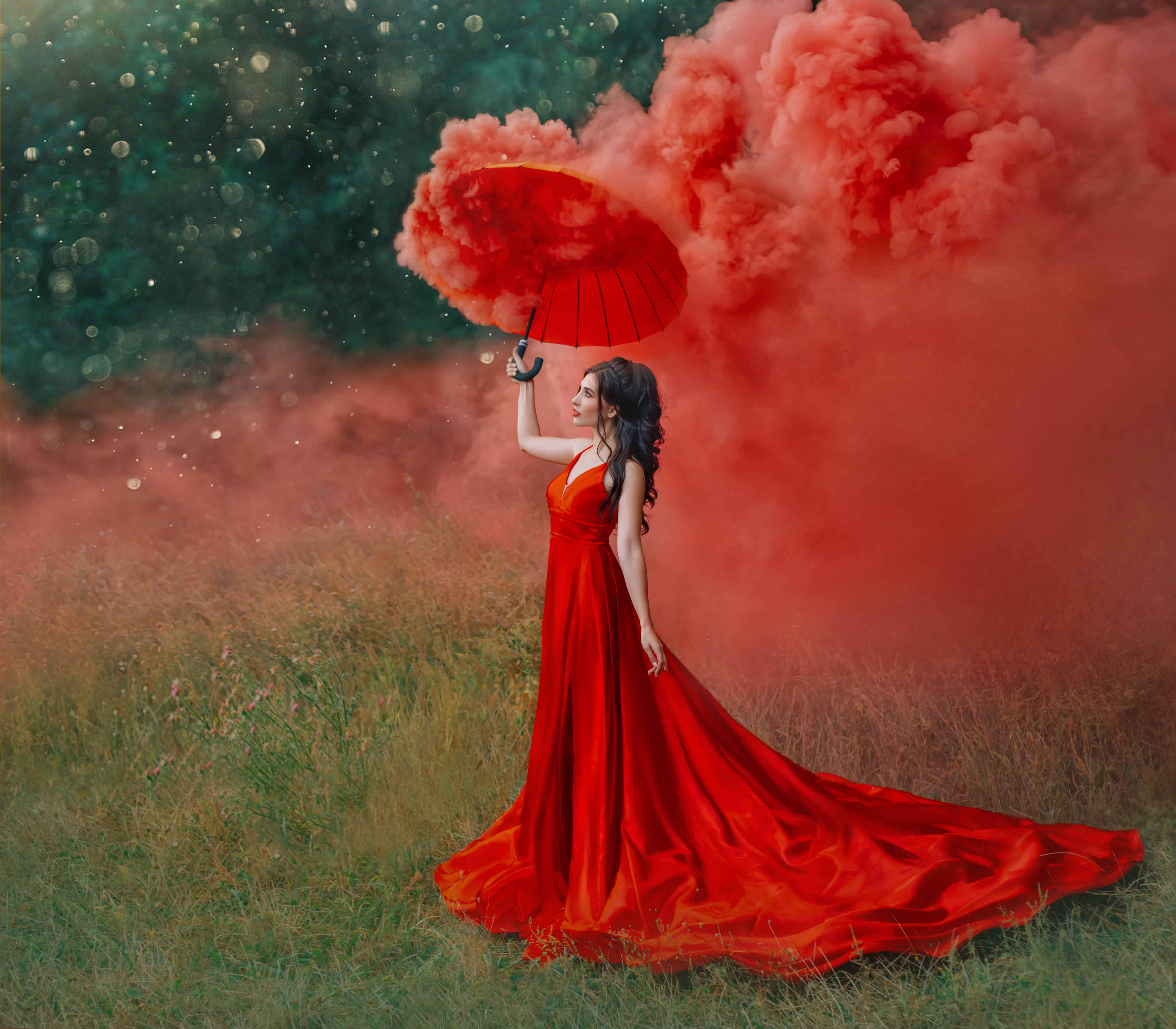 woman in red silk long dress train with umbrella. Art design photography. Idea Creative photo shoot with colored smoke bomb. Magical light nature. Glamorous fashion lady walks in woods. valentines day