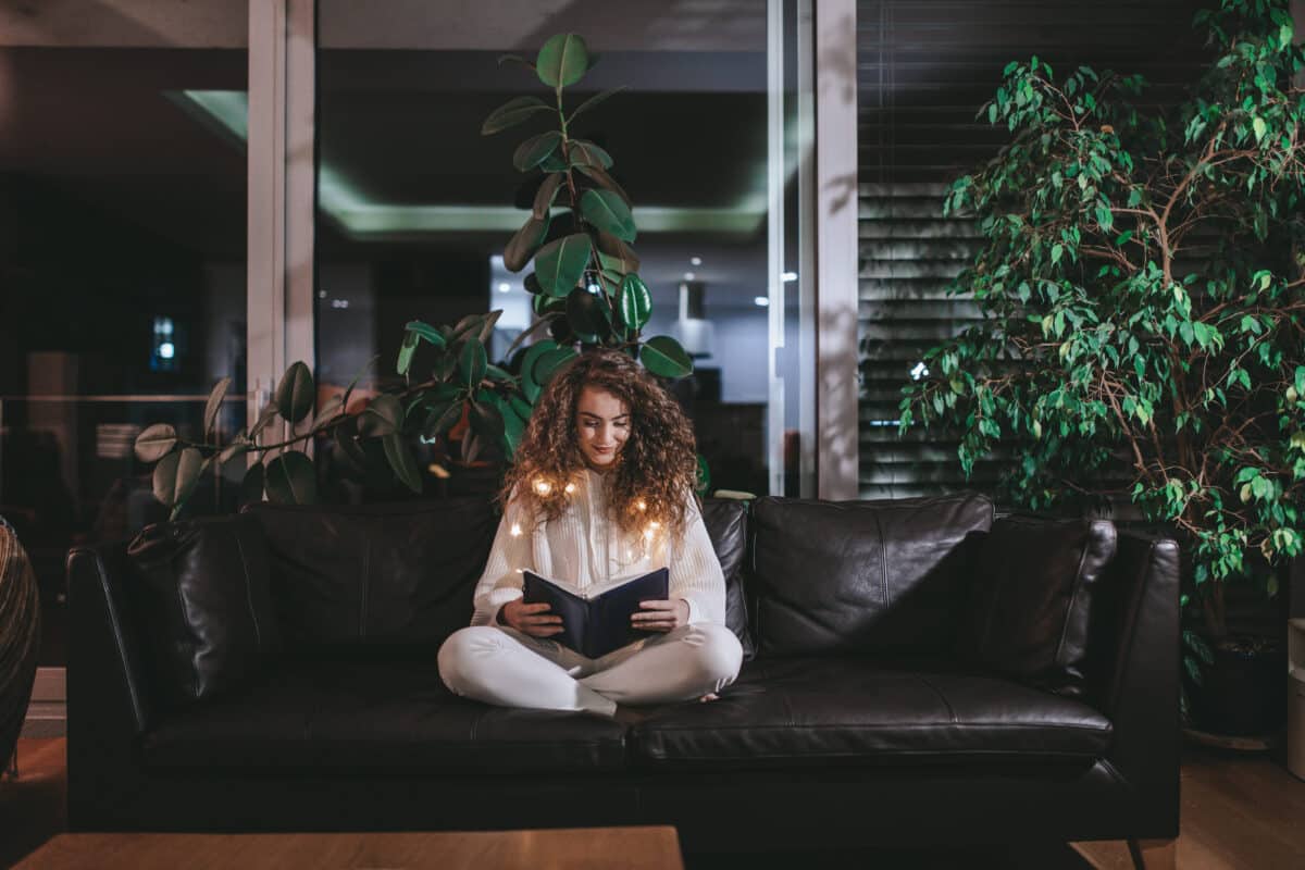 Happy young woman sitting on sofa and reading book in the evening in cozy hyyge living room.