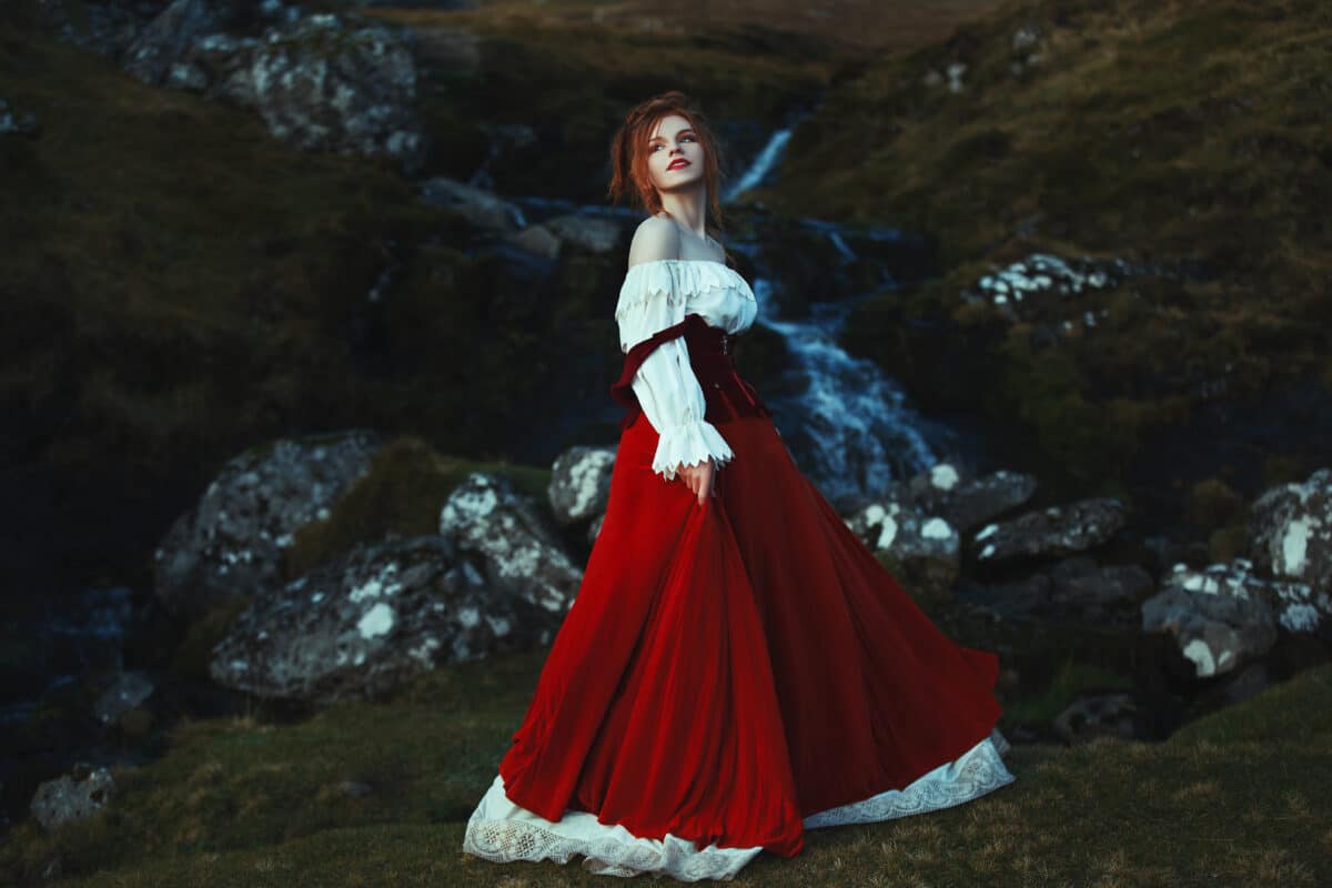 Beautiful red-haired girl dressed white and red costume is smiling on the background of stones covered with moss and waterfall