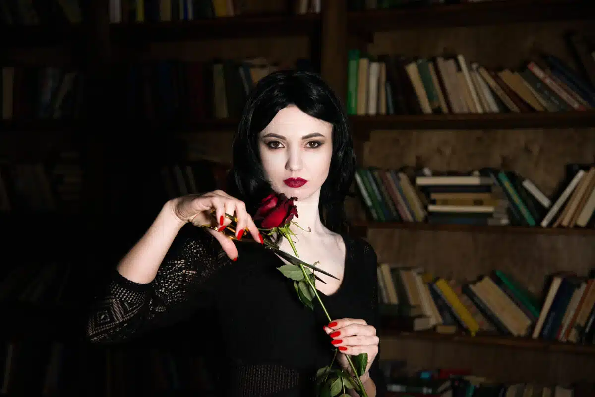 beautiful brunette girl cosplay morticia addams with red lips and nails holding red rose and scissors