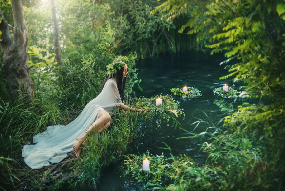 Fabulous mythical natural landscape. Forest fantasy woman sits on shore lake, nymph throws wreath in water. Old Slavic cult ceremony Ivan Kupala. Long white dress. Black hair