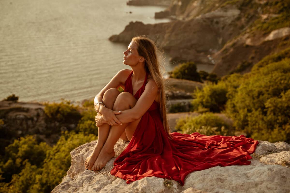 Sea woman sunset coast. Beautiful sensual woman in a long red dress and with long hair, sitting on a rock above the beautiful sea in a large bay.