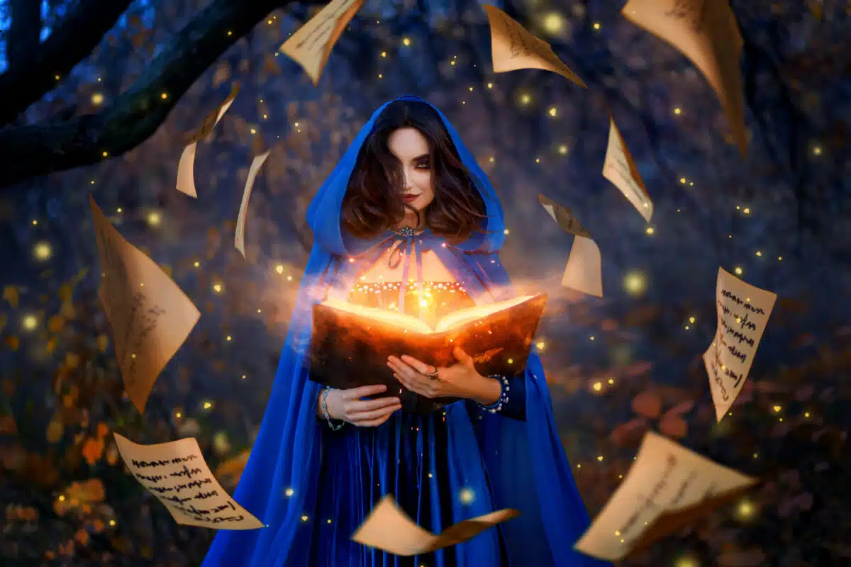 Fantasy woman witch magician in hood holds in hands magic book, bright orange light spells, wind scatters fall sheets paper page levitation. Girl sorceress. Medieval cloak blue dress magician costume.