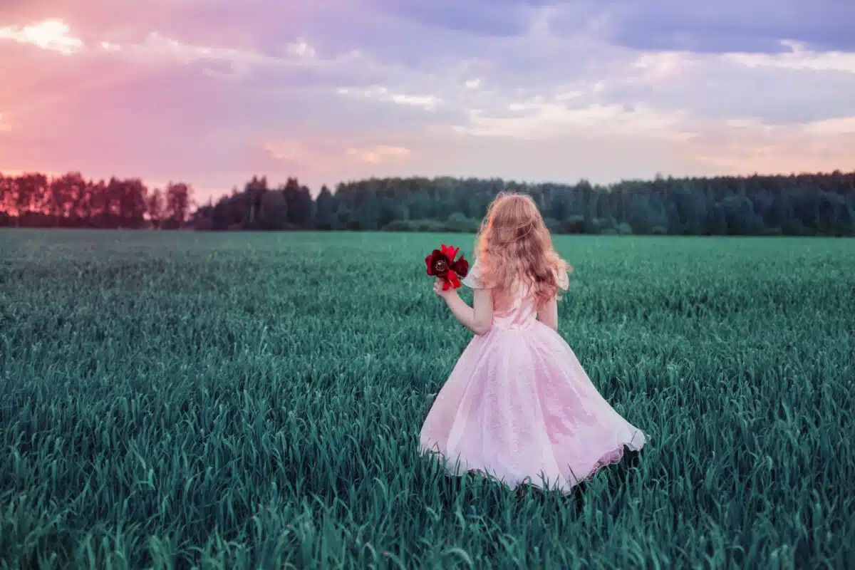 princess in a pink dress is walking in the green field holding a red poppy flower
