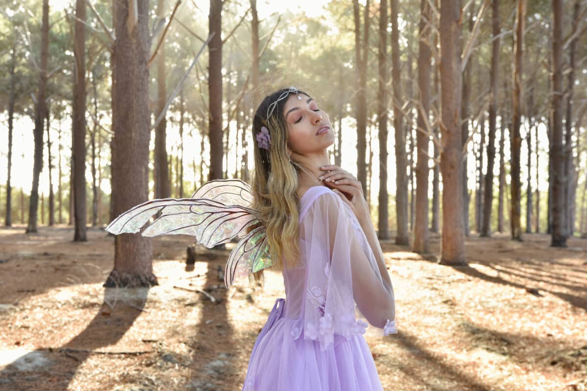 portrait of beautiful young blonde model wearing a purple princess fantasy ball gown with flower crown diadem, posing with butterfly wings. pine tree forest location background with golden lighting