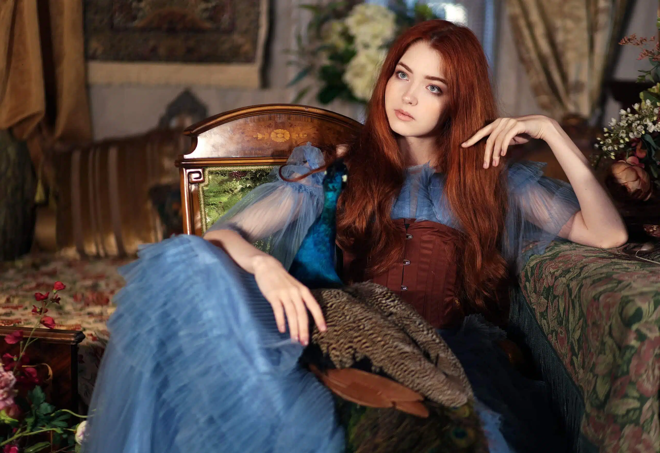 Young red haired woman in long blue dress holding a blue peacock