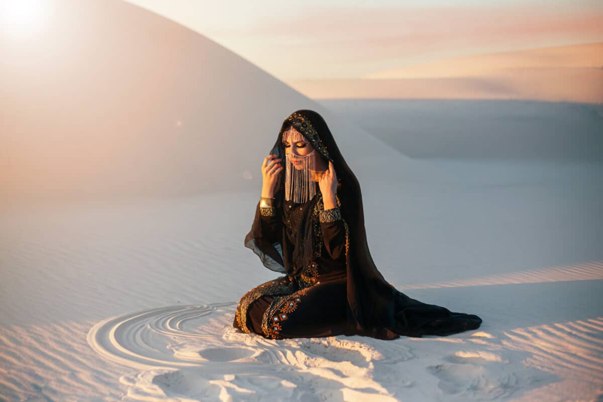 A mysterious woman in a black long dress sits in the desert. Luxurious clothes, gold accessories hide the face. Oriental beauty fashion model. Sand dunes background, orange sunset . Art Fantasy photo