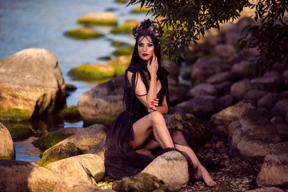 Caucasian Brunette Female Ballerina In Black Dress Posing on Rocky Shore While Wearing Seashell Decorated Crown And Long Black Pearl Necklace.