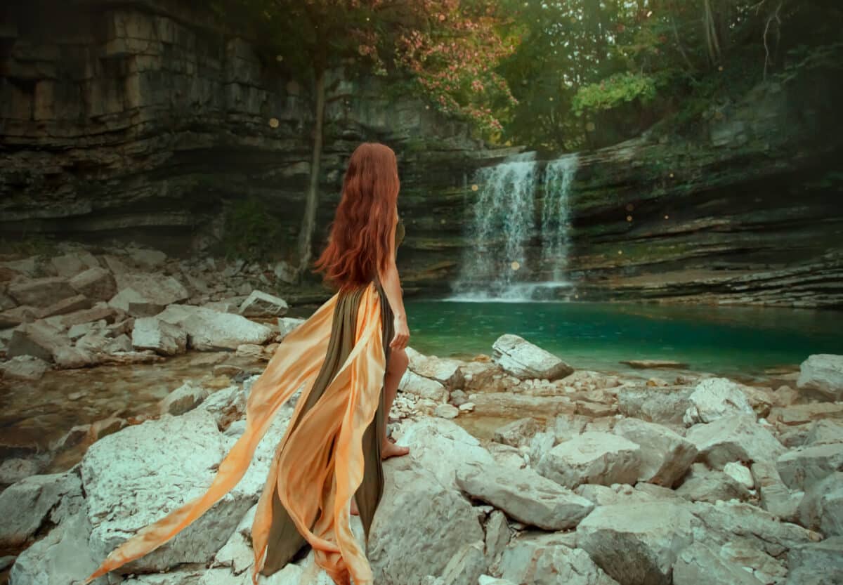 Slim redhead girl in a green dress, with a waving train, stands with his back to the camera. The princess is looking at the waterfall lagoon. Without a face. Art photo of a river mermaid
