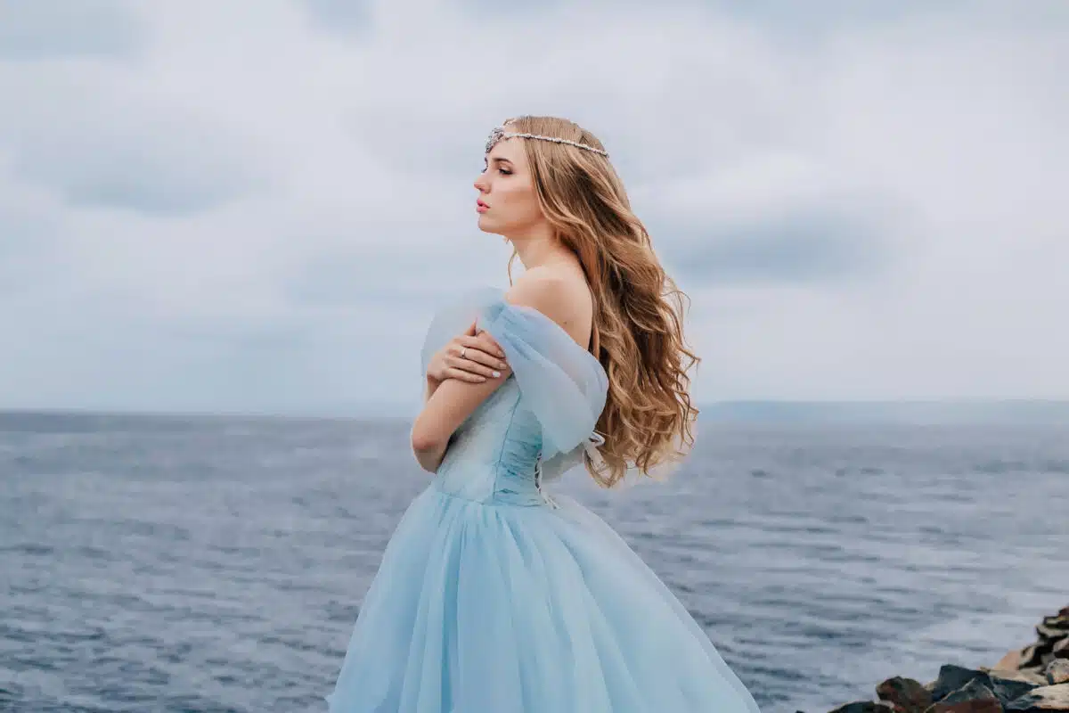 Beautiful young girl in a long blue lush dress is standing on the beach.