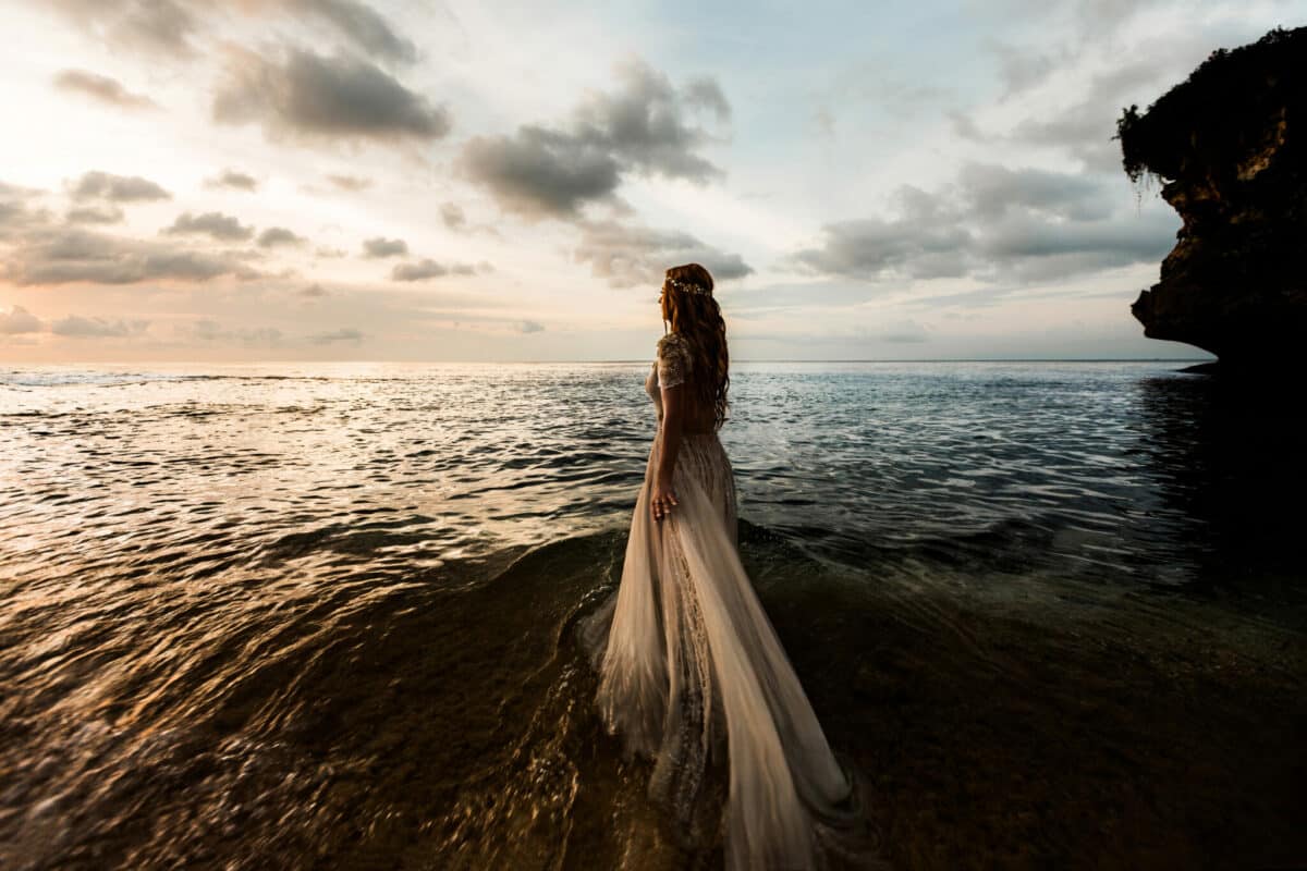 A bride in a white wedding dress on the beach admires the sunset, standing in the sea water.