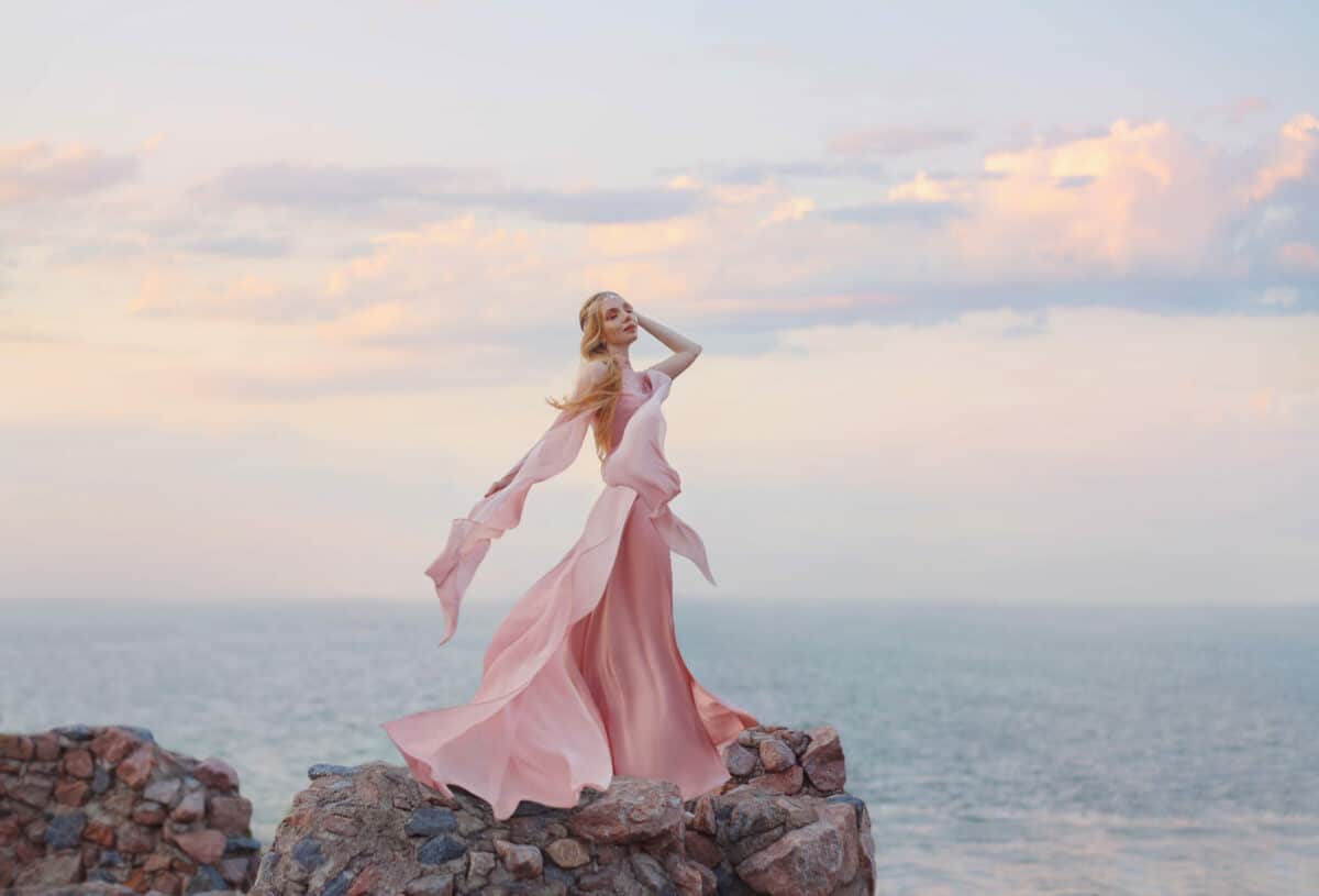 elegant girl elf with blond fair wavy hair with tiara on it, wearing a long light pink rose rozy fluttering dress, standing on the high tower of the old castle.