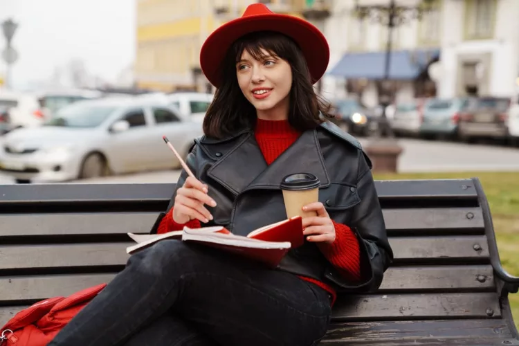 pretty young woman wearing leather jacket and red hat writing while seated on a bench outdoor.